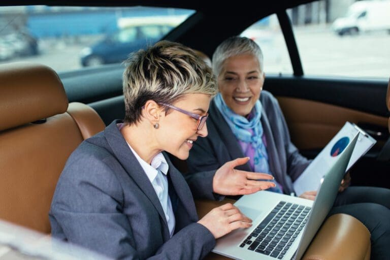 Two professional women discussing work on a laptop inside a car.